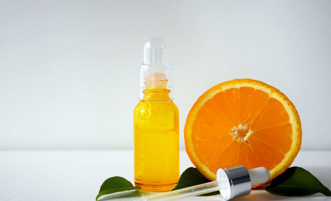 Serum z witaminą C w butelce, a obok niego owoc pomarańczy.