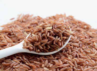 Ryż naturalny brązowy rozsypany na białym blacie. Na nim biała łyżeczka z ryżem.